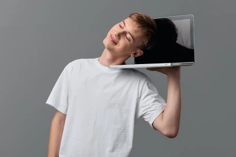 Man in a white t shirt rests a laptop on his shoulder, eyes closed in a relaxed pose, conveying tech fatigue, casual indoor scene, balance and creative break