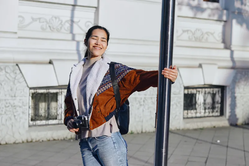 Smiling woman on an outdoor street with camera, casual jacket and scarf, leaning on a pole in daylight, urban ambiance and candid style for lifestyle photography and street capture vibes