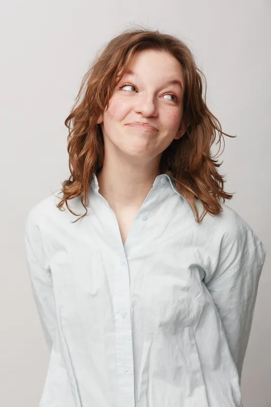 Young woman with curly hair wearing white shirt looks sideways with playful smile. Studio portrait on light background. Concept of casual style, happiness, and natural expression.