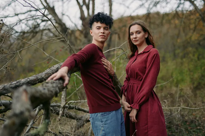 Authentic, candid portrait of a young couple outdoors, wearing burgundy outfits, standing by a rustic fence amid bare branches, conveying real emotion and genuine connection.