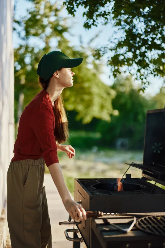 Young woman grilling outdoors in a green cap and casual attire, enjoying a sunny day in a natural setting with vibrant greenery