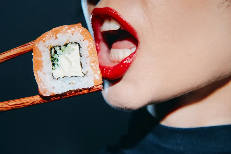 Крупный план ярко-красных губ женщины, которая наслаждается вкусным роллом с суши, подчеркивающий свежесть суши на фоне драматичного темного фона