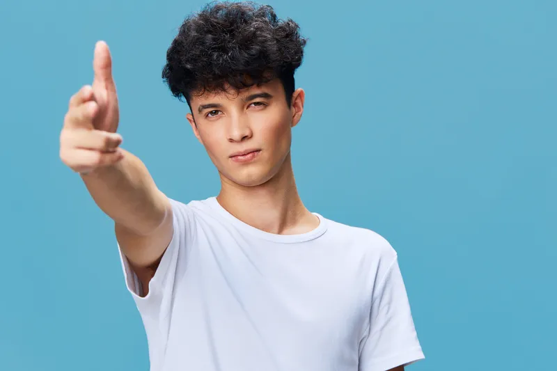 young man in white t-shirt pointing finger with confident expression against blue background, casual style, studio portrait, focused attitude