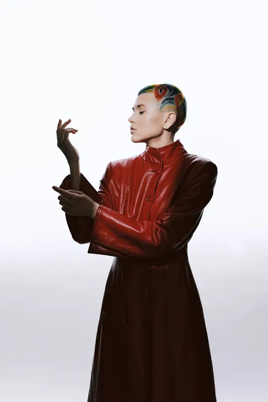 Woman with colorful buzz cut hairstyle wearing a stylish red leather coat, posing confidently against a minimalist white background with soft lighting.
