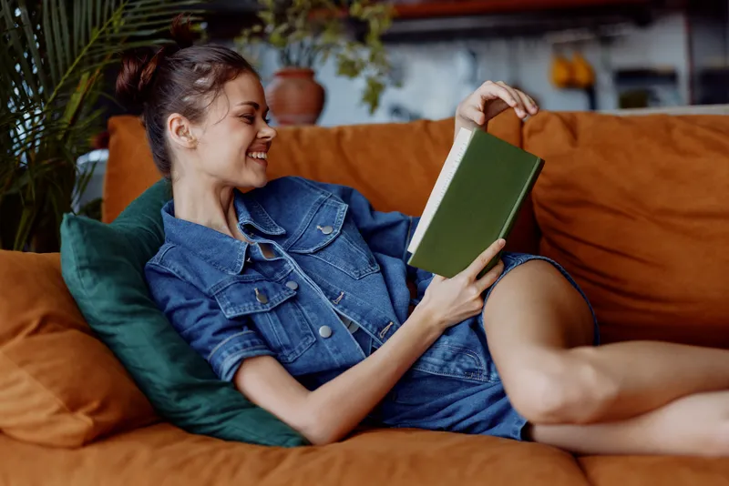 Молодая женщина отдыхает на диване, читает книгу в уютной гостиной с комнатным растением в горшке