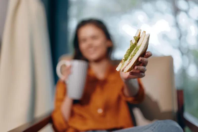 Весёлая молодая женщина наслаждается вкусным тостом с авокадо, держа в руке чашку кофе