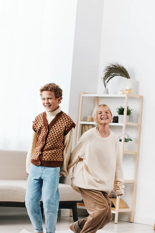 Two joyful children laughing and dancing indoors in a bright modern living space, wearing cozy casual outfits, capturing playful energy and carefree happiness.