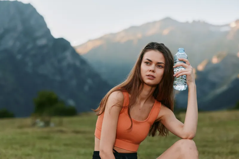 A young outdoor woman sits in a serene valley beside mountains, holding a water bottle and looking toward the camera with calm confidence and intent gaze.