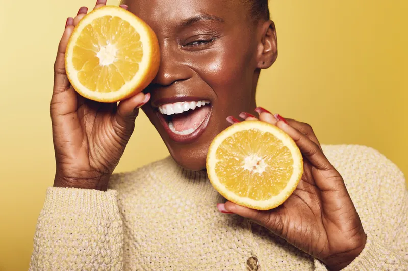 Happy African woman with dark skin holds two halves of fresh orange, smiling with closed eyes against yellow background. Concept of healthy lifestyle, natural beauty, and vitamin C.