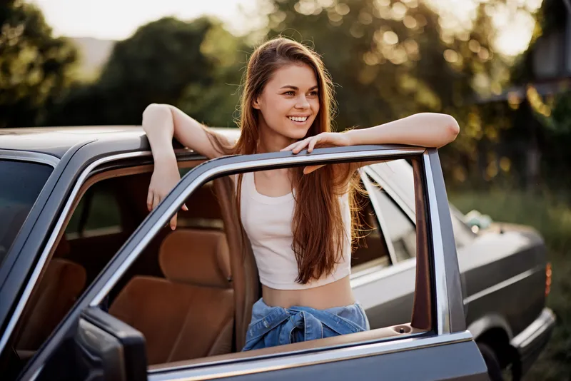 A happy young woman leaning on a car door during sunset, showcasing casual summer fashion in a vibrant outdoor setting, evoking a sense of freedom and adventure.