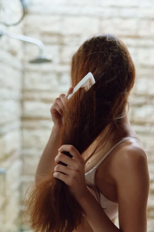 Woman combing long hair in a bright bathroom with tiled walls, casual white tank top, natural light highlighting soft strands and texture.