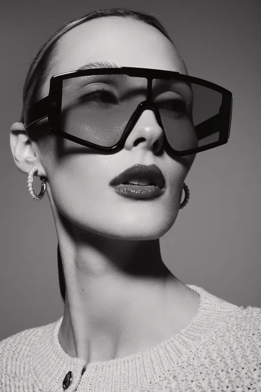 Portrait of a white woman with pale skin wearing oversized sunglasses and hoop earrings in black and white studio lighting.
