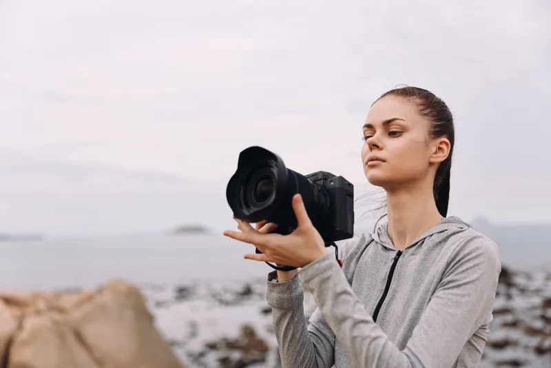 Young woman with a camera on a rocky beach, focused and determined in a gray hoodie under a cloudy sky, capturing nature s beauty during a serene moment