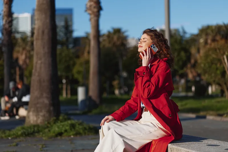 Smiling woman in a red coat sits outdoors in a sunny park, talking on a phone. Casual lifestyle in an urban setting, warm natural light, relaxed mood.