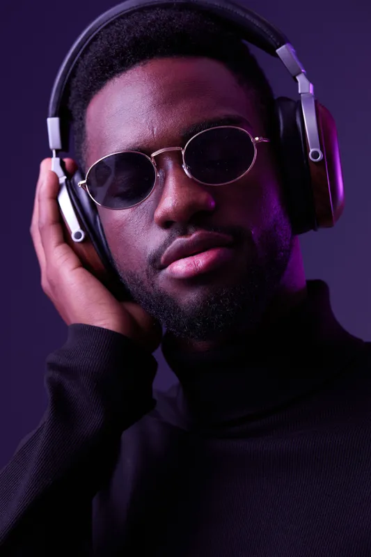 Stylish young man with headphones, wearing sunglasses, enjoying music in a modern setting with a vibrant purple background, expressing relaxation and confidence