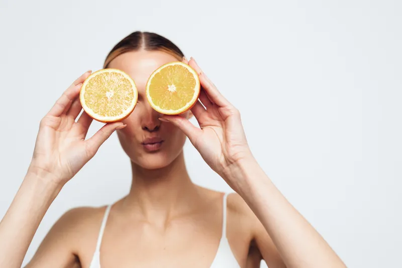 Playful young woman holds two halved oranges in front of her eyes, creating a bright whimsical portrait in a clean studio with soft lighting and a white background.