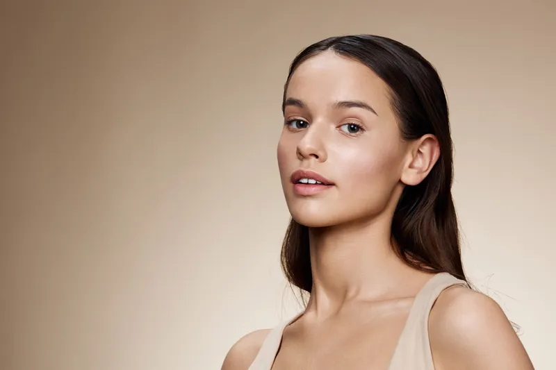 Elegant young model with natural makeup and long hair, set against a warm beige background, expressing confidence and beauty in a minimalistic style