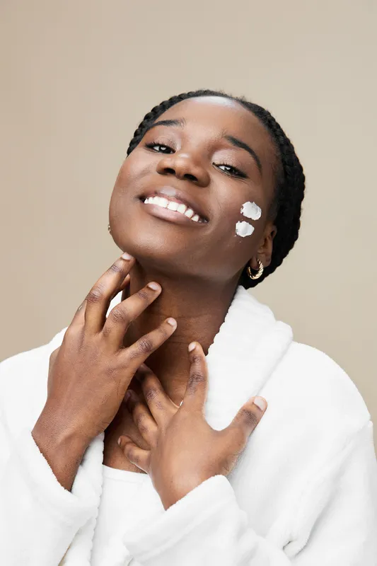 Smiling young woman enjoying skincare routine with face cream, showcasing glowing skin and joyful expression against a neutral beige background