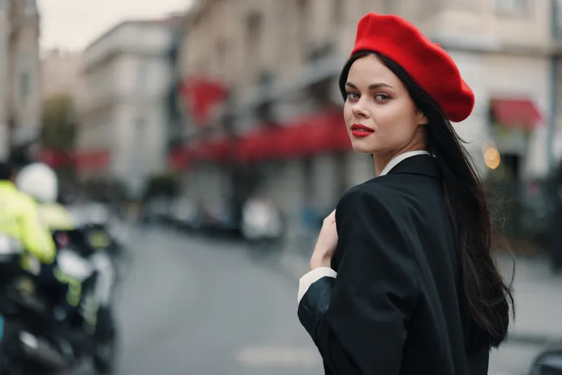 Портрет модной женщины-красавицы, стоящей на улице на фоне города в стильной одежде, с красными губами и красным беретом