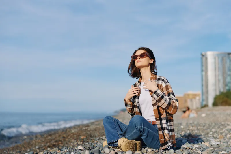 Woman sitting on a pebble beach by the sea, wearing sunglasses and a plaid shirt, casual style, relaxed mood with ocean breeze and clear sky horizon ahead