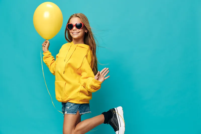Young girl in yellow hoodie and red heart sunglasses holding a yellow balloon with cheerful smile, posing on blue background in casual summer outfit and black sneakers.