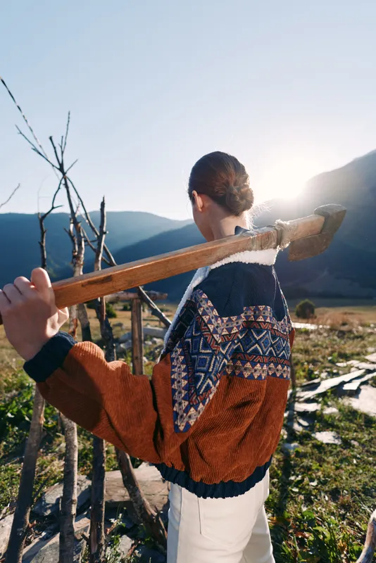 Woman with axe on shoulder in rural mountains field at sunrise, wearing patterned sweater and white pants, walking by wooden fence on a farm under bright sunlight and calm scenery.