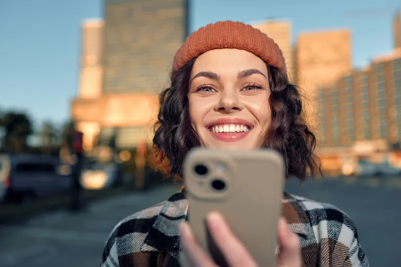 Woman smile smartphone portrait in urban scene at golden hour, candid beanie look conveying authenticity and mindful living, emotional storytelling with sincere modern city lifestyle.