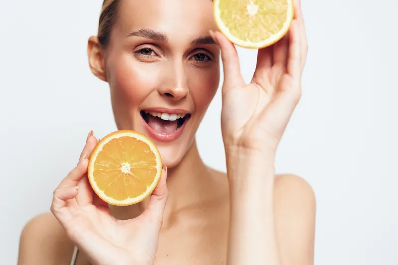 Smiling woman holding two orange halves near her face on a white background. Concept of healthy lifestyle, freshness, and natural vitamins. Bright studio lighting highlights skin texture.