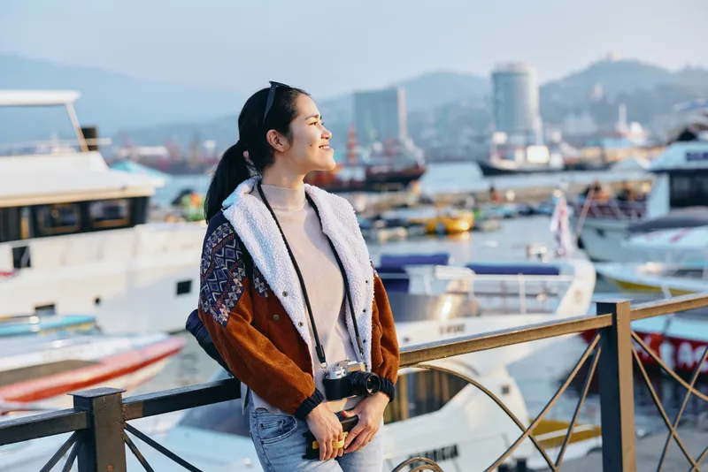 Woman by harbor boats waterfront smiling wearing stylish coat enjoying outdoor fashion lifestyle by marina promenade daylight scene with breeze water glow cozy casual scenic urban breeze horizon