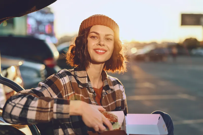 woman smile pizza outdoors sunset beanie urban candid. Young woman eats pizza from a box in a parking lot at golden hour, authenticity and candid lifestyle with joyful mindful living vibe.