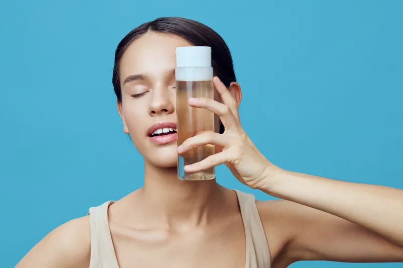 Young woman holding a bottle of skincare product against a blue background, expressing relaxation and confidence, embodying healthy skin and beauty