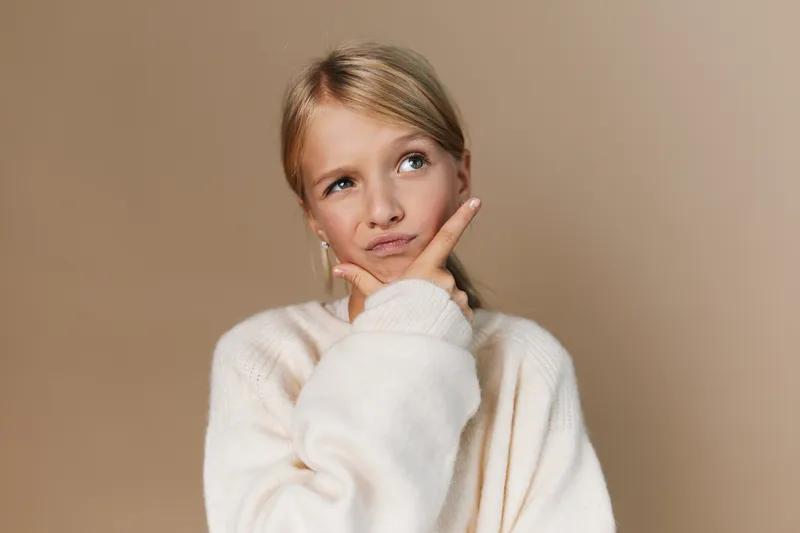 A thoughtful young girl wearing a soft white sweater stands against a warm beige backdrop, her finger resting on her chin as she studies a gentle, pensive mood and curious gaze, inviting quiet