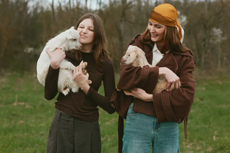 Two women hold young goats in a green field, enjoying a carefree rural moment. A candid lifestyle scene celebrating friendship, animals, and outdoor living in nature.