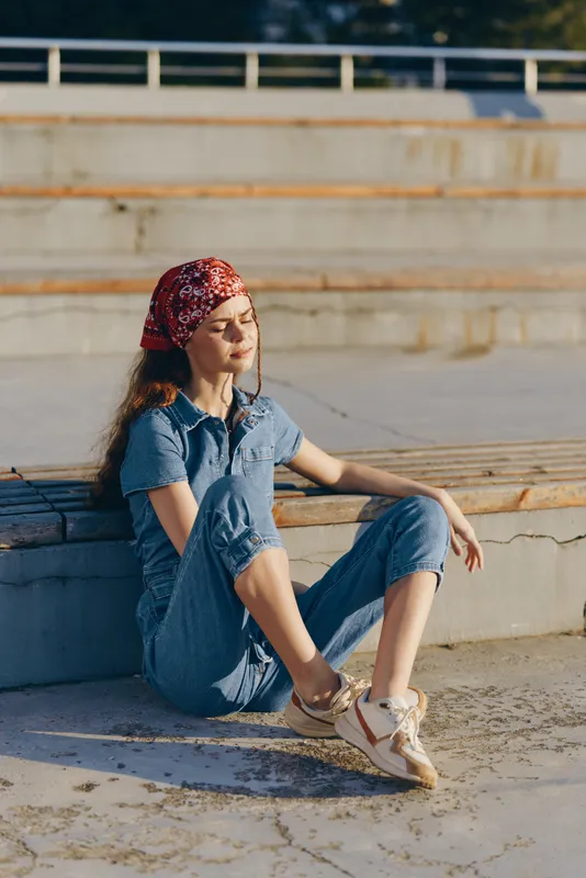 Relaxed woman in modern boho-western denim street style sitting outdoors in warm sunlight, wearing casual sneakers and a red bandana, captured with film color effect.
