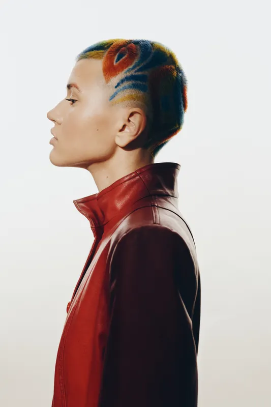 Woman with colorful shaved hairstyle in profile wearing a red leather jacket against a bright white background, showcasing modern and edgy style.