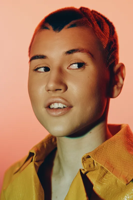 Young woman with braided hair and warm brown skin wearing a yellow shirt, looking to the side against a soft peach background under warm lighting.