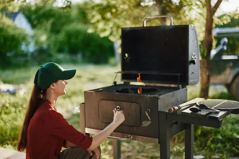 Young woman grilling outdoors in a green summer setting, wearing a baseball cap and a relaxed outfit, enjoying the warm weather and vibrant atmosphere