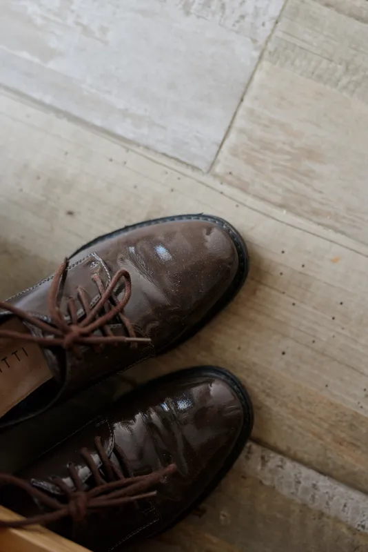 Casual home scene shows brown leather shoes resting on a wooden floor, suggesting a quiet morning routine and relaxed domestic atmosphere with subtle hints of everyday life around