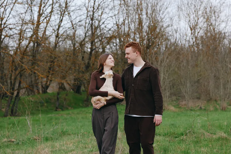 A couple walks outdoors in a park with a small dog, sharing smiles and gentle affection, casual clothing, bright greenery around, candid moment of connection and companionship