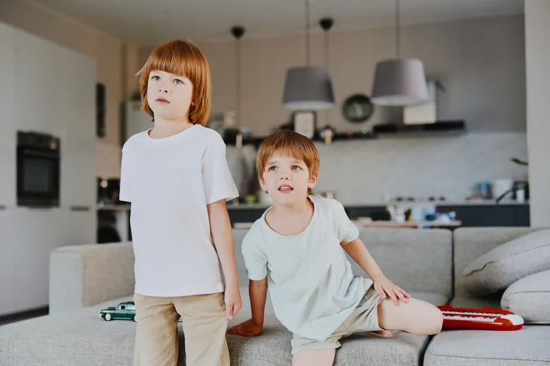 Two children in casual clothing standing in a cozy living room, showcasing curiosity and playfulness. The warm ambiance enhances their cheerful expressions.