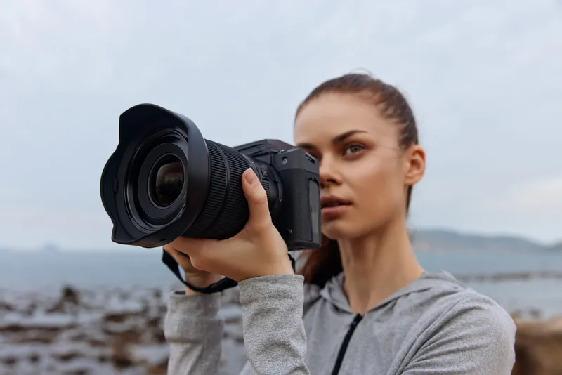 Young woman photographing a scenic landscape with a professional camera in a gray hoodie near the ocean, showcasing creativity and passion for photography