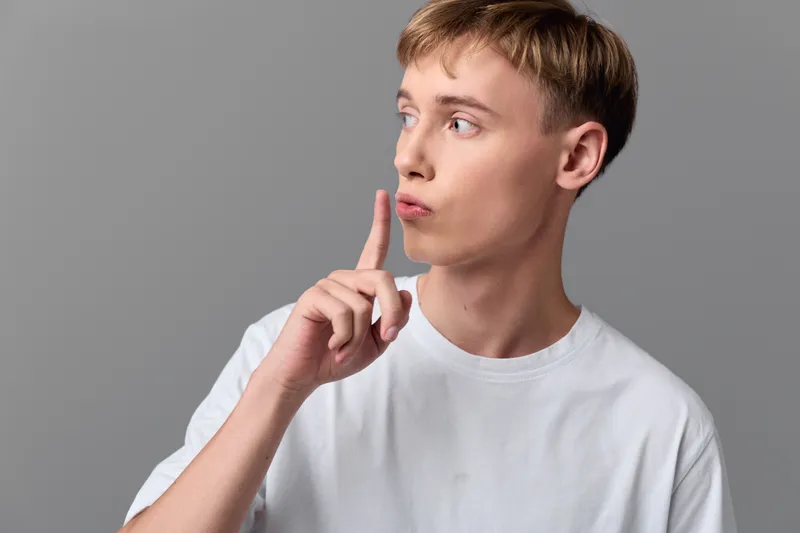 Shushing gesture by a man in a white tee, finger to lips signaling secrecy, hush, or quiet moment, calm restraint and discreet communication for creative projects or film