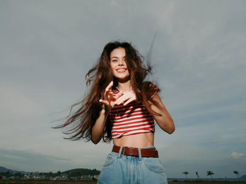 A joyful young woman dances outdoors under a clear blue sky, wearing a striped crop top and denim jeans with a brown belt, hair flowing, bright smile, and energetic summer vibe.