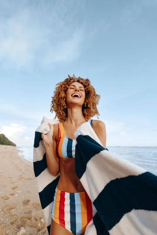 Young woman on a sunny beach wears a vibrant striped swimsuit and wraps in a towel, smiling with joy as warm waves softly touch the shore.