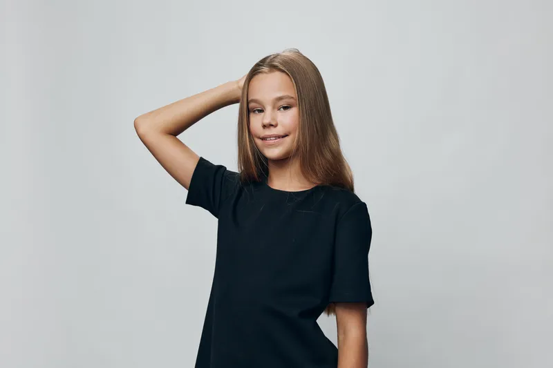 Girl with long hair wearing black t shirt posing with hand on head smiling against white background in studio. Casual child portrait with confident expression and relaxed posture.