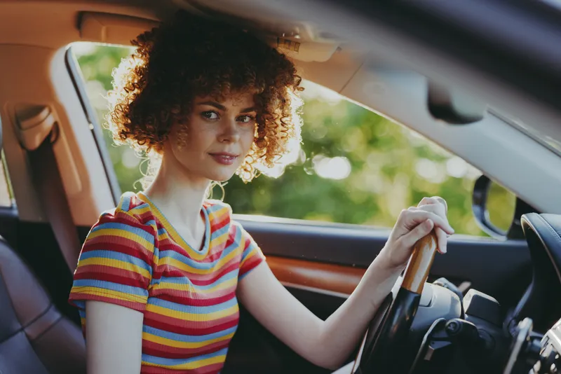 Young woman with curly hair sits in a colorful striped shirt behind the wheel, captured in a warm retro film tone, in a car with soft natural light and a confident look on a quiet street at golden