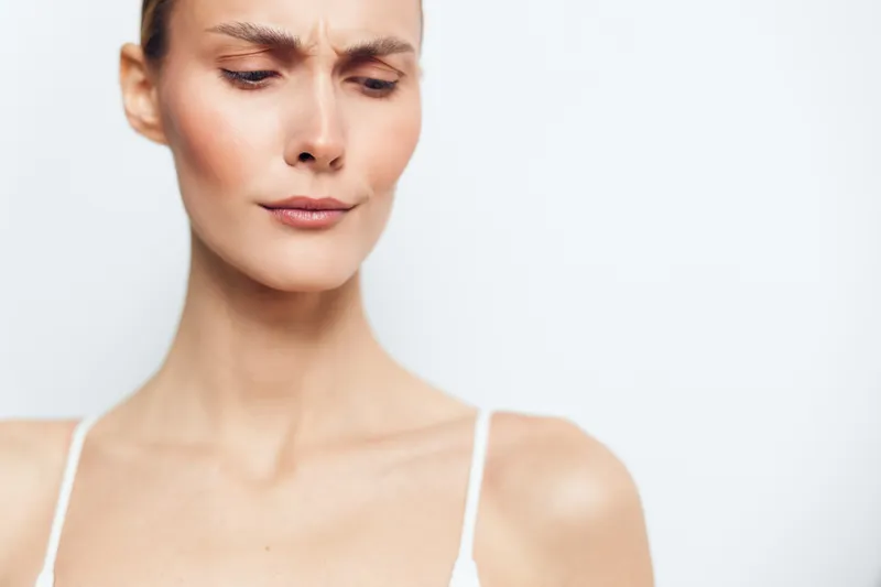 Young woman with clear skin looking down with a serious expression on a white background. Concept of natural beauty, skincare, and minimalism with soft natural lighting.
