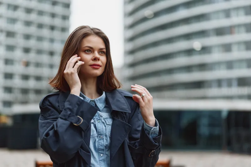 Young woman in a denim shirt and dark trench talks on a phone in an urban courtyard, confident, focused, with modern glass buildings behind her.