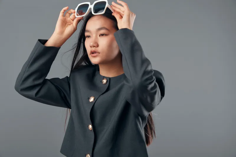 Woman posing with sunglasses in hands against gray background wearing modern black jacket with buttons. Young female model with long hair styling her glasses for casual fashion shoot or portrait.