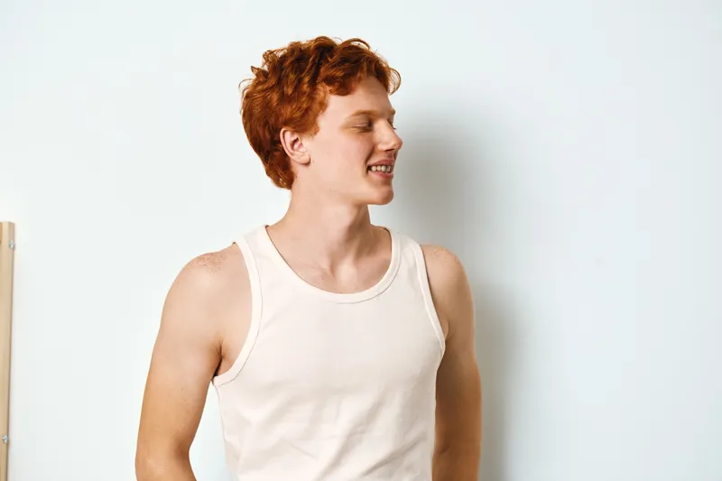 Young man with red hair wearing white sleeveless shirt standing against white wall smiling and looking sideways in casual modern style indoors with natural light.